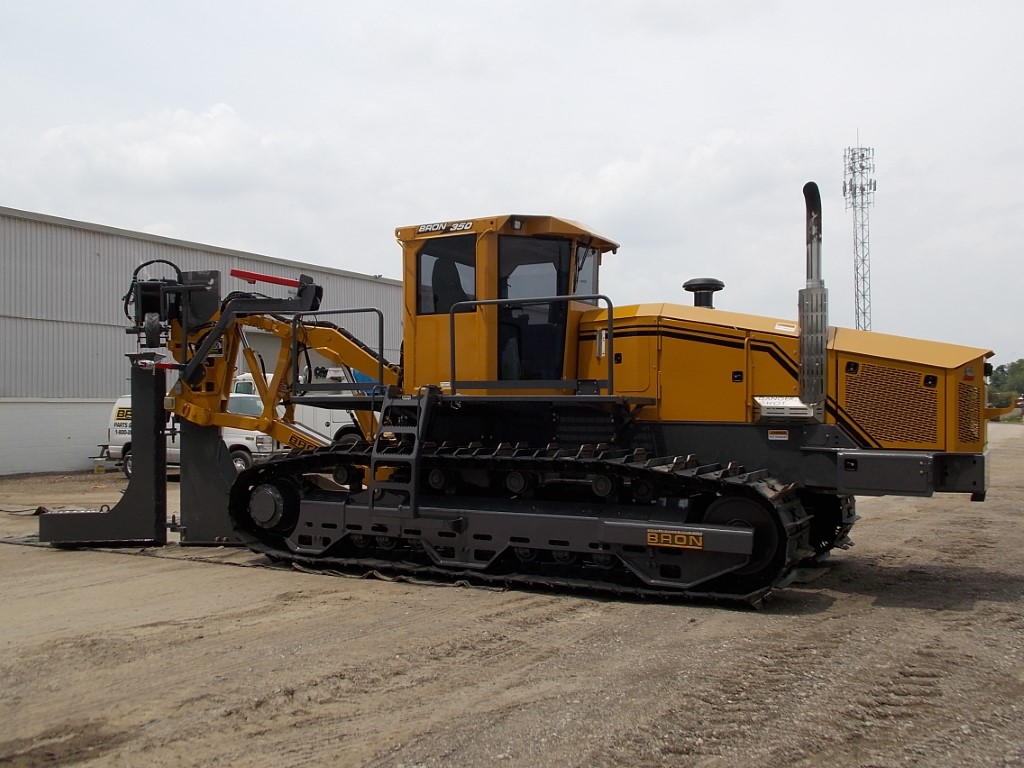 BRON utility tractor with yellow and black coloring, featuring a robust plow and cable installation equipment, parked on a gravel lot in front of an industrial warehouse.