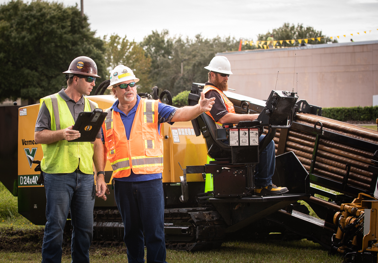 Three Vermeer Southeast employees in safety gear, including helmets and reflective vests, at a construction site. Two are discussing project details over a tablet, while the third operates a horizontal directional drill in the background.