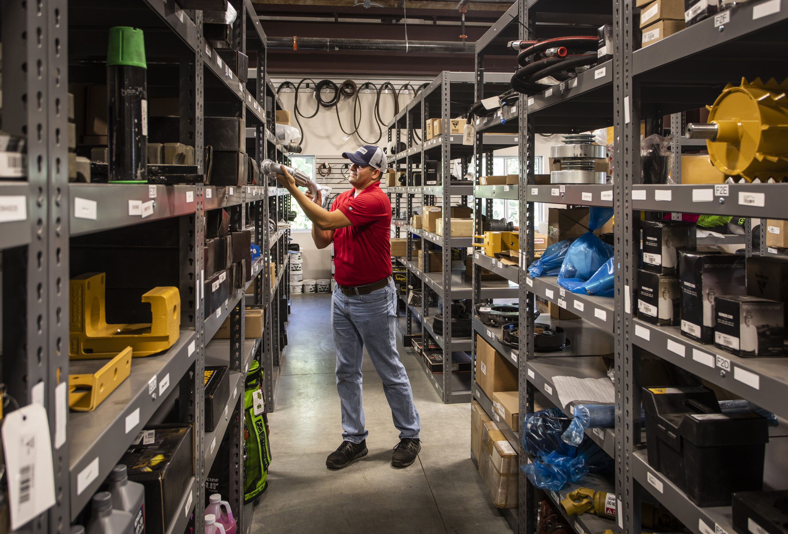 A male employee in a red shirt is retrieving parts from a well-organized, stocked shelf in the Vermeer Southeast parts warehouse. The shelves are filled with various heavy equipment parts and accessories.