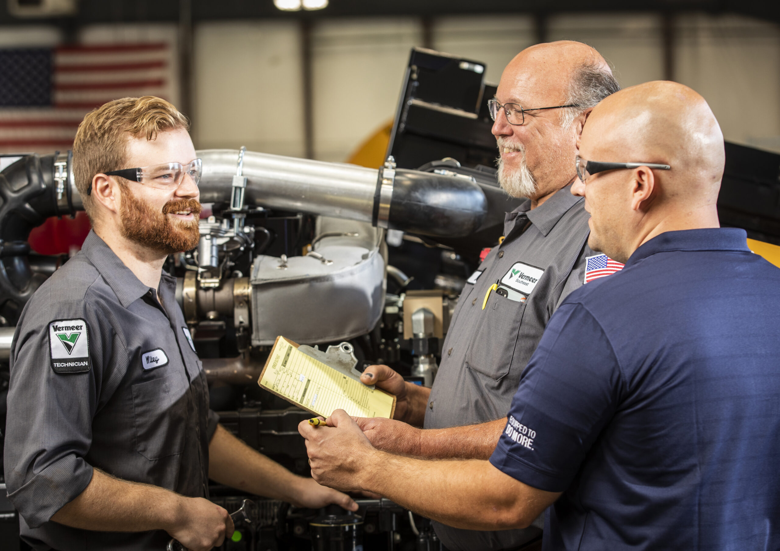 Three male Vermeer Southeast employees, two technicians and the Service Director, wearing company uniforms, engaging in a discussion over a manual in the workshop. They are standing in front of a large piece of heavy machinery, smiling and collaborating on maintenance work.