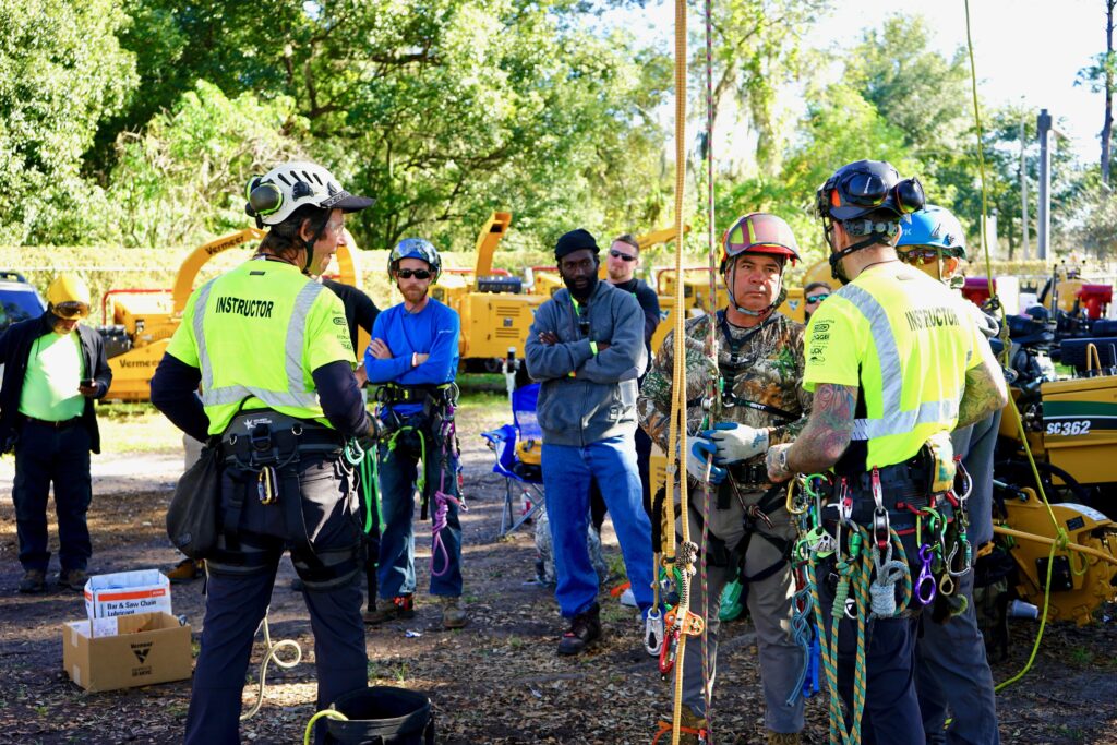 Instructors and participants at a Vermeer arborist training event discuss equipment and techniques. The group, wearing climbing gear and safety helmets, stands near heavy machinery in an outdoor setting with trees in the background.