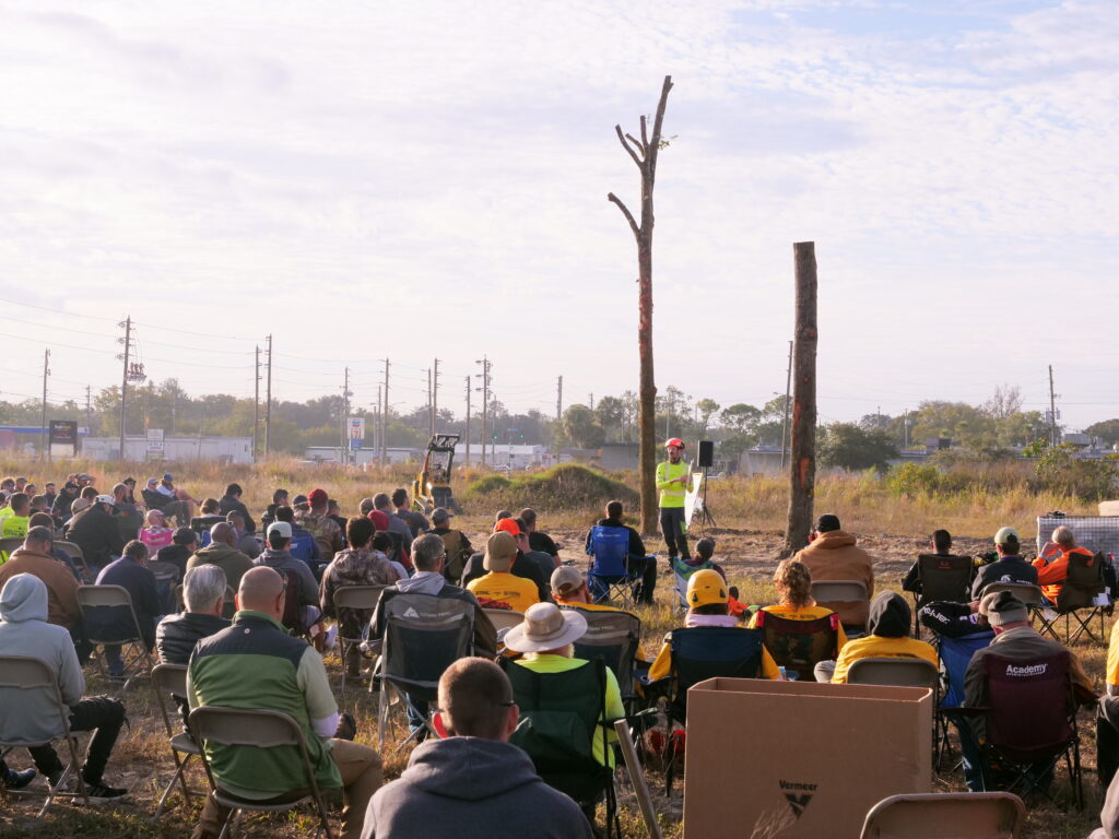 Attendees at a Vermeer outdoor arborist training session watch a trainer demonstrate tree climbing on a bare trunk. The group, in safety gear, sits in folding chairs in a field under a clear sky with distant power lines visible.