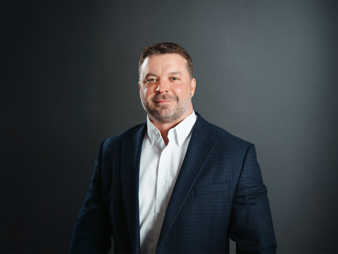 Professional studio portrait of a man wearing a dark blazer over a white button-down shirt against a dark background.