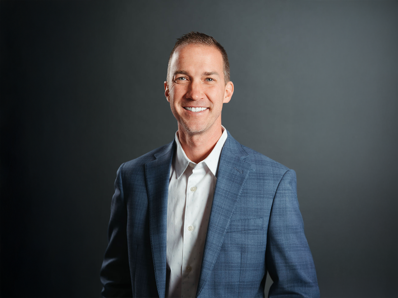 Professional studio headshot of a person wearing a blue blazer over a white button-down shirt, standing against a neutral gray background.